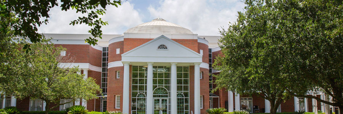 Florida State University’s College of Law building with a domed roof, tall white columns, large glass entryway, and red brick exterior, framed by trees and greenery.