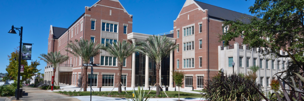 Florida State University’s Legacy Hall, a large brick academic building with tall windows, palm trees, and a central columned entrance, shown under a bright blue sky.